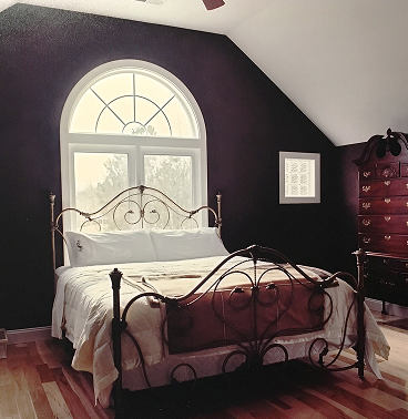 A bed with white sheets up against a black wall with a white-rimmed window in the middle